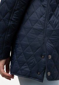 Námořnický prošívaný kabát s hladkou, lesklou texturou. Výrazné rysy zahrnují diamantový vzor a dva černé knoflíky na zapínání vpředu.