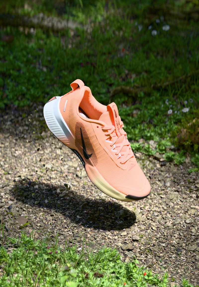 Lekka buty do biegania w pastelowym pomarańczowym kolorze z teksturowaną cholewką, białą podeszwą, czarnym logo Swoosh oraz wyraźnym systemem sznurowania.