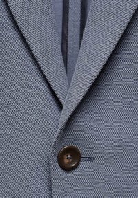 Gros plan d'un blazer gris texturé avec un revers cranté et un seul bouton marron foncé fermé à l'avant.