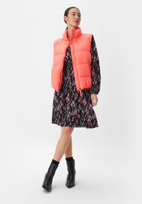 Padded, bezrękawnik w jasnym koralowym kolorze z wysokim kołnierzem, noszony na czarnej sukience z kolorowym wzorem geometrycznym. Czarne botki sięgające do kostek.