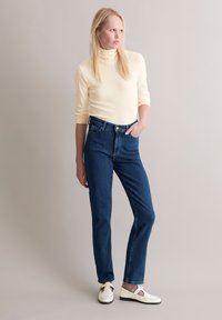 Haut à col roulé jaune clair, jean en denim bleu foncé taille haute et chaussures blanches avec un design ajouré. Silhouette simple et ajustée.