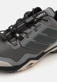 Sive tenisice za sport s teksturiranim mrežastim gornjim dijelom, oblikovanim preklopima, crnim vezicama, reflektirajućim detaljima i čvrstim gumenim potplatom.