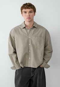 Kariertes Hemd in Braun- und Cremetönen, mit einer einzelnen Brusttasche und lässiger Passform, kombiniert mit dunkelgrauen Hosen.