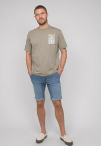 T-shirt en coton vert olive à manches courtes avec une poche graphique sur le devant, associé à un short en jean et des chaussures à enfiler beiges.