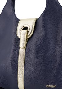 Marine lædertaske med en tekstureret guldaccentrem og rund hardwaredetalje, med en glat overflade og diskret branding.