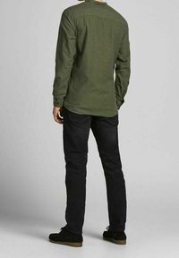 Man i en olivgrön långärmad skjorta, svarta jeans och svarta skor, stående med ryggen mot en neutral bakgrund.