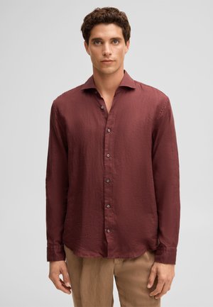 Młody mężczyzna w bordowej koszuli zapinanej na guziki z długim rękawem i beżowych spodniach, stojący na jednolitym jasnoszarym tle.