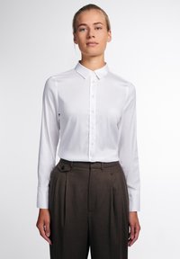 Camicia bianca con colletto classico, maniche lunghe, tessuto morbido e vestibilità su misura abbinata a pantaloni marroni a vita alta.