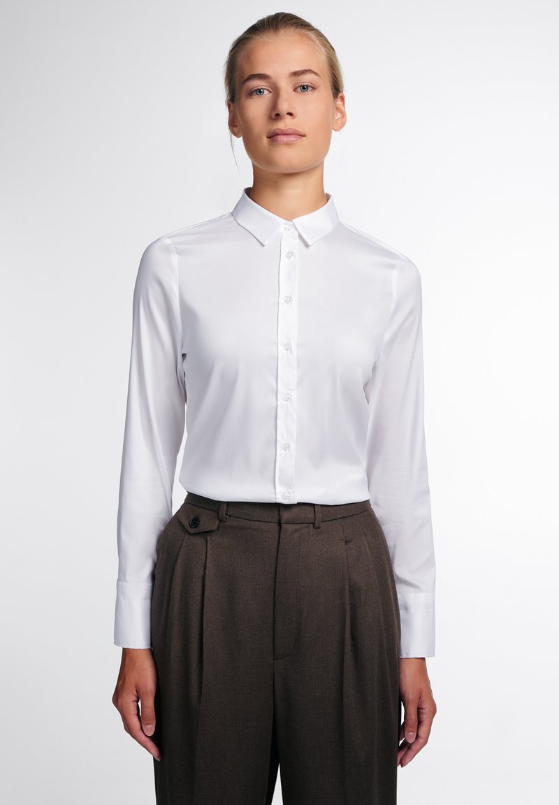 Camicia bianca con colletto classico, maniche lunghe, tessuto morbido e vestibilità su misura abbinata a pantaloni marroni a vita alta.