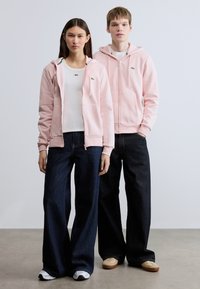 Ροζ φούτερ με φερμουάρ και μπροστινές τσέπες, φορεμένο πάνω από λευκό τοπ, συνδυασμένο με σκούρο μπλε παντελόνι φαρδιάς γραμμής. Τα αθλητικά παπούτσια ολοκληρώνουν το σύνολο.