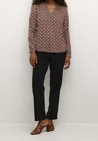 Langarmbluse mit V-Ausschnitt, die mit einem kreisförmigen Blumenmuster in Pink-, Creme- und dunklen Tönen verziert ist, kombiniert mit schwarzen Hosen und Absatzsandalen.
