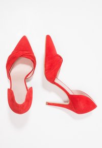 Chaussures à talons hauts en daim rouge avec un bout pointu, un design noué à l'avant et un dos ouvert. Caractéristiques : talon fin et surface texturée.
