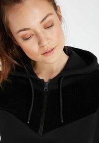 Zwarte zip-up hoodie met een gedetailleerd corduroy paneel op de bovenborst, verstelbare trekkoorden en een ontspannen pasvorm ontwerp.
