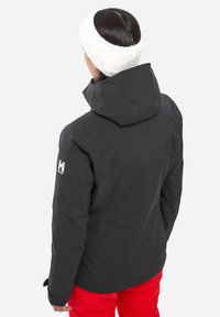 Personne aux cheveux sombres portant une veste à capuche noire, un pantalon rouge et un bandeau blanc, tourné le dos contre un fond blanc.