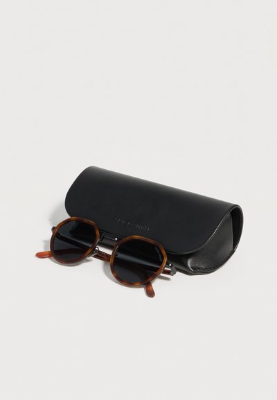 Στρογγυλά καφέ γυαλιά ηλίου με σχέδιο κοχύλι χελώνας και σκούρους φακούς δίπλα σε μια μαύρη κυρτή θήκη σε λευκό φόντο.