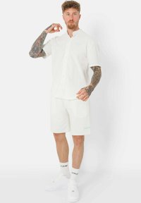Chemise blanche à manches courtes avec un devant à boutons, assortie à un short. Présente un texte bleu clair et des baskets blanches.