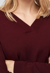 Blonde Person trägt einen bordeauxroten Pullover mit V-Ausschnitt, die Arme verschränkt, zeigt den oberen Brustbereich und einen Teil der Ärmel.