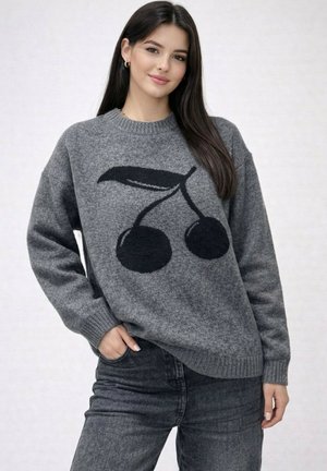 Jeune femme aux longs cheveux foncés portant un pull gris avec un motif de cerise noire et un jean gris foncé, debout, une main dans la poche.