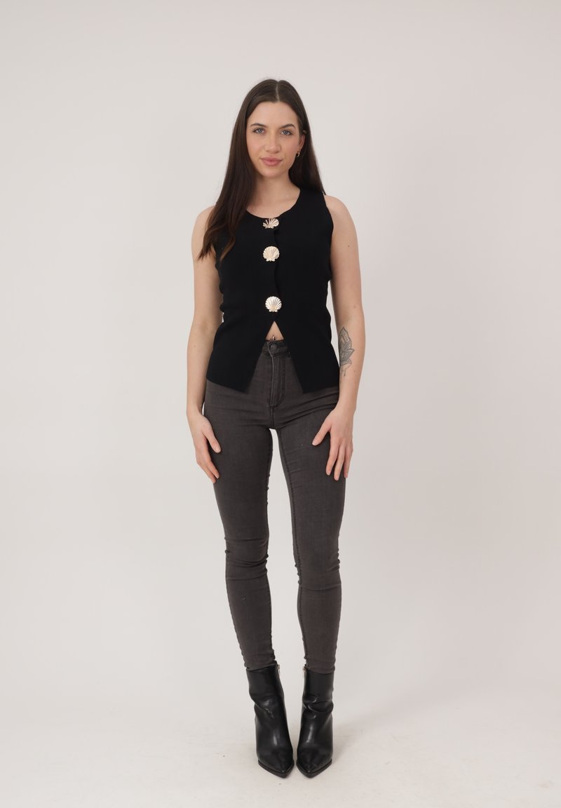 Frau steht und trägt ein ärmelloses schwarzes Top mit großen Muschelknöpfen, dunkle Skinny-Jeans, schwarze Stiefeletten und hat ein Tattoo am linken Unterarm.