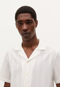 Camisa blanca de manga corta con cuello abotonado, que presenta rayas verticales y una textura sutil, y botones de color amarillo claro.