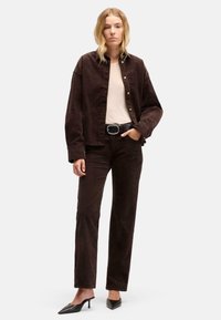 Giacca in velluto a coste marrone e pantaloni abbinati, indossati con una maglia chiara e una cintura nera. Presenta una vestibilità comoda e tacchi neri a punta.