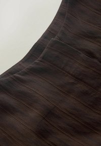 Detail tmavě hnědé látky s jemnými vertikálními pruhy a detaily šití, zobrazující texturu a překrývající se vrstvy.