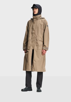 Mand iført en beige lang regnfrakke med knap- og lynlåslukning, sort hætte og bukser samt sorte sko, stående mod en ensfarvet baggrund.