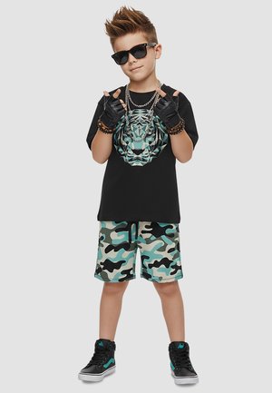 Junge mit Sonnenbrille, schwarzen Handschuhen, schwarzem Tiger-Print-Shirt, Camouflage-Shorts und schwarzen Sneakers, der eine Handbewegung macht.