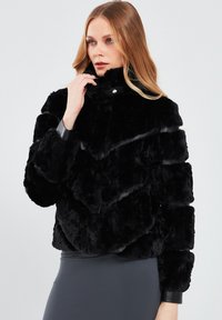 Black faux fur jacket with a high collar, chevron pattern, and glossy black leather accents on the cuffs and neckline.