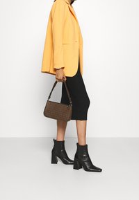 Gelber Blazer, schwarzes geripptes Kleid und schwarze Stiefeletten. Hält eine braune Handtasche mit Muster und kurzem Griff.