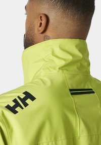 Chaqueta impermeable de un brillante color amarillo, con un alto cuello, que presenta acentos de logotipo en negro y un diseño elegante. Textura suave y corte moderno.
