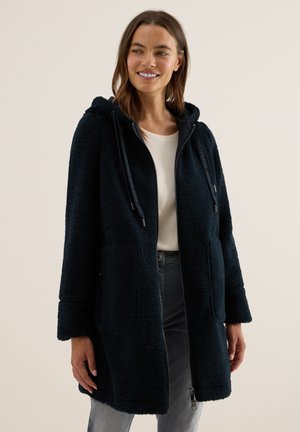 Femme portant un long manteau en sherpa noir zippé avec capuche, se tenant contre un fond clair, souriante et regardant sur le côté.