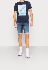 Laivastonsininen t-paita, jossa on ananasgrafiikka ja "Jack & Jones" -teksti, yhdistettynä vaaleansinisiin farkkushortseihin, joissa on käännetyt lahkeet, ja valkoisiin tennareihin.