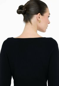 Donna con capelli scuri raccolti in un chignon intrecciato, che indossa un maglione a coste nero e orecchini ovali dorati, di profilo su uno sfondo bianco.