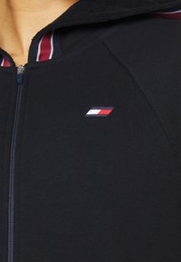 Felpa con zip nera a coste. Presenta un piccolo logo ricamato in rosso, bianco e blu sul lato sinistro del petto. Dettagli a strisce nel colletto.