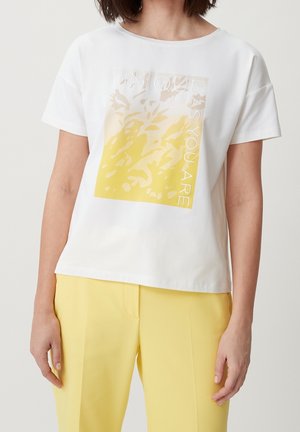 Femme portant un T-shirt blanc à manches courtes avec un motif floral jaune et beige et le texte « UNIQUE AS YOU ARE », assorti à un pantalon jaune.
