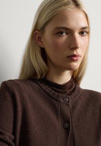 Bruine gebreide cardigan met geribbelde halslijn en twee knopen, gedragen over een bijpassende turtleneck. Stevige textuur, zachte stof, minimalistisch ontwerp.