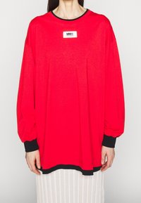 Person som bär en överdimensionerad långärmad tröja i klar röd färg med svarta muddar och nederkant, som har en vit rektangulär logotypapplikation på bröstet.