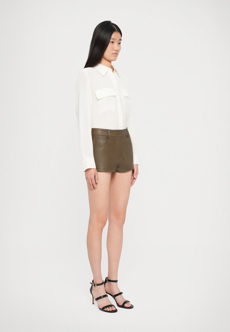 Camisa blanca de manga larga con dos bolsillos en el frente, combinada con pantalones cortos de cuero verde oliva de cintura alta y sandalias negras de tacón alto con tiras.