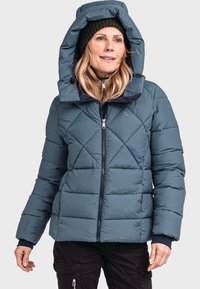 Padded türkisfarbener Jacke mit großer Kapuze, Reißverschluss vorne und Rautenmotiv; getragen über einem schwarzen Shirt und einer Hose aus strukturiertem Stoff.