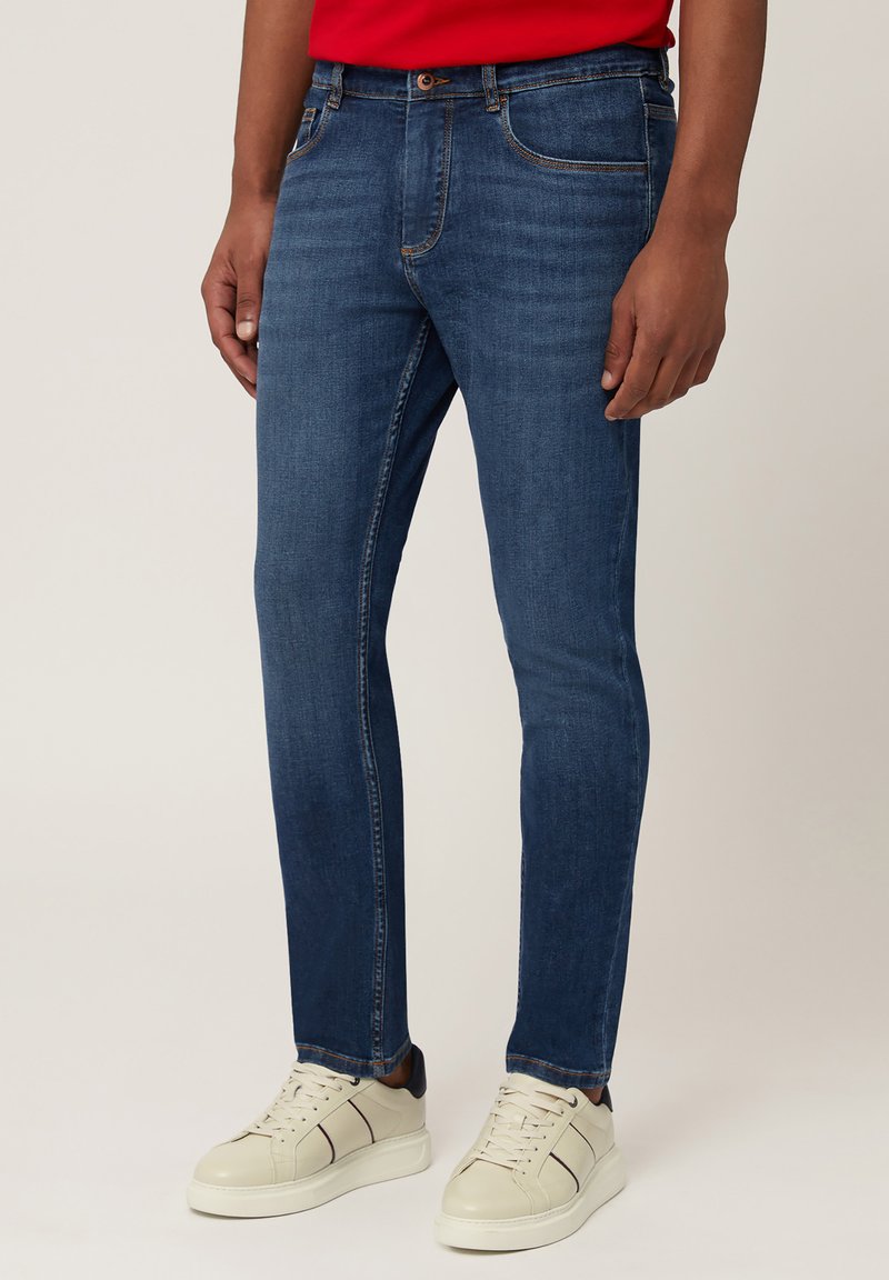 Slim-fit modri denim kavbojke s svetlo pranjem, ki imajo pet žepov in kontrastno šivanje, v kombinaciji z belimi športnimi copati.