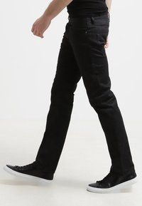 Vaqueros de denim negro con un corte ajustado, que presentan una textura suave, diseño de cinco bolsillos y herrajes metálicos, combinados con zapatillas negras.