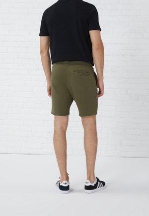 Olivgrüne Shorts mit glatter Textur, elastischem Bund und einer Gesäßtasche. Kombiniert mit einem schwarzen T-Shirt und schwarz gestreiften Schuhen.
