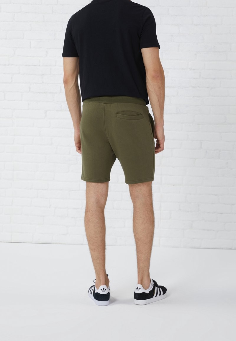 Shorts vert olive avec une texture lisse, une taille élastique et une poche arrière. Associés à un t-shirt noir et des chaussures noires à rayures.
