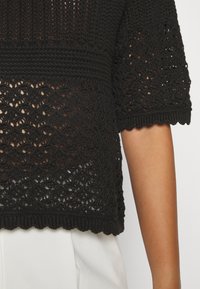 Svart virkad topp med öppna vävmönster, korta ärmar, texturerade detaljer och en vågig fåll. Plagget har en avslappnad passform.