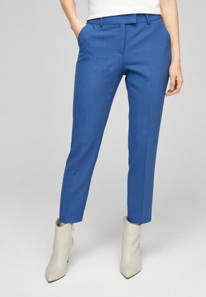 Vrouw draagt op maat gemaakte blauwe enkellange broek en gebroken-witte enkellaarsjes met puntige neus, staand tegen een effen achtergrond.