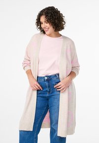 Gai beigu adītu kardigānu ar rozā sirds rakstiem, atvērta priekšpuse, garas piedurknes un ribu malām, uzliktu virs rozā kreklam un ziliem džinsiem.