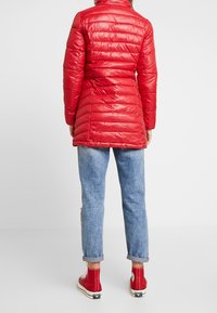 Chaqueta de abrigo roja con costuras horizontales, mangas largas y cuello alto, llevada sobre unos vaqueros ligeros azul claro arremangados y zapatillas rojas de caña alta.