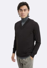 Maglione nero lavorato a maglia con scollo a V, maniche lunghe e polsini a costine. Indossato sopra un dolcevita grigio, con pantaloni grigi a motivo.