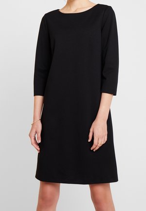 Femme portant une robe noire simple longueur genoux avec des manches trois-quarts, debout devant un fond gris clair uni.